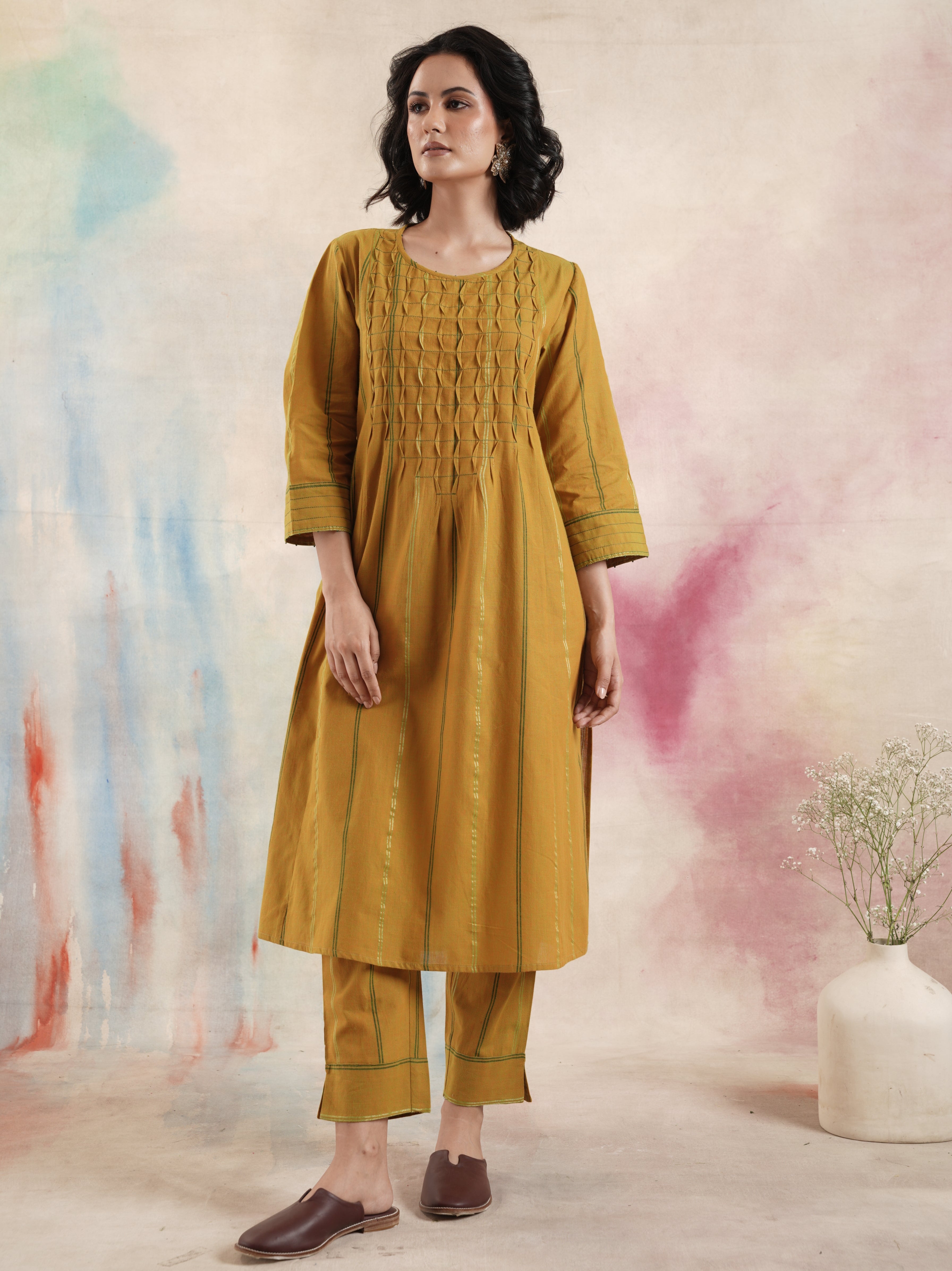 A woman standing against a colorful backdrop, wearing a mustard yellow pure mangalgiri kurta set with ruching and hand bead work.