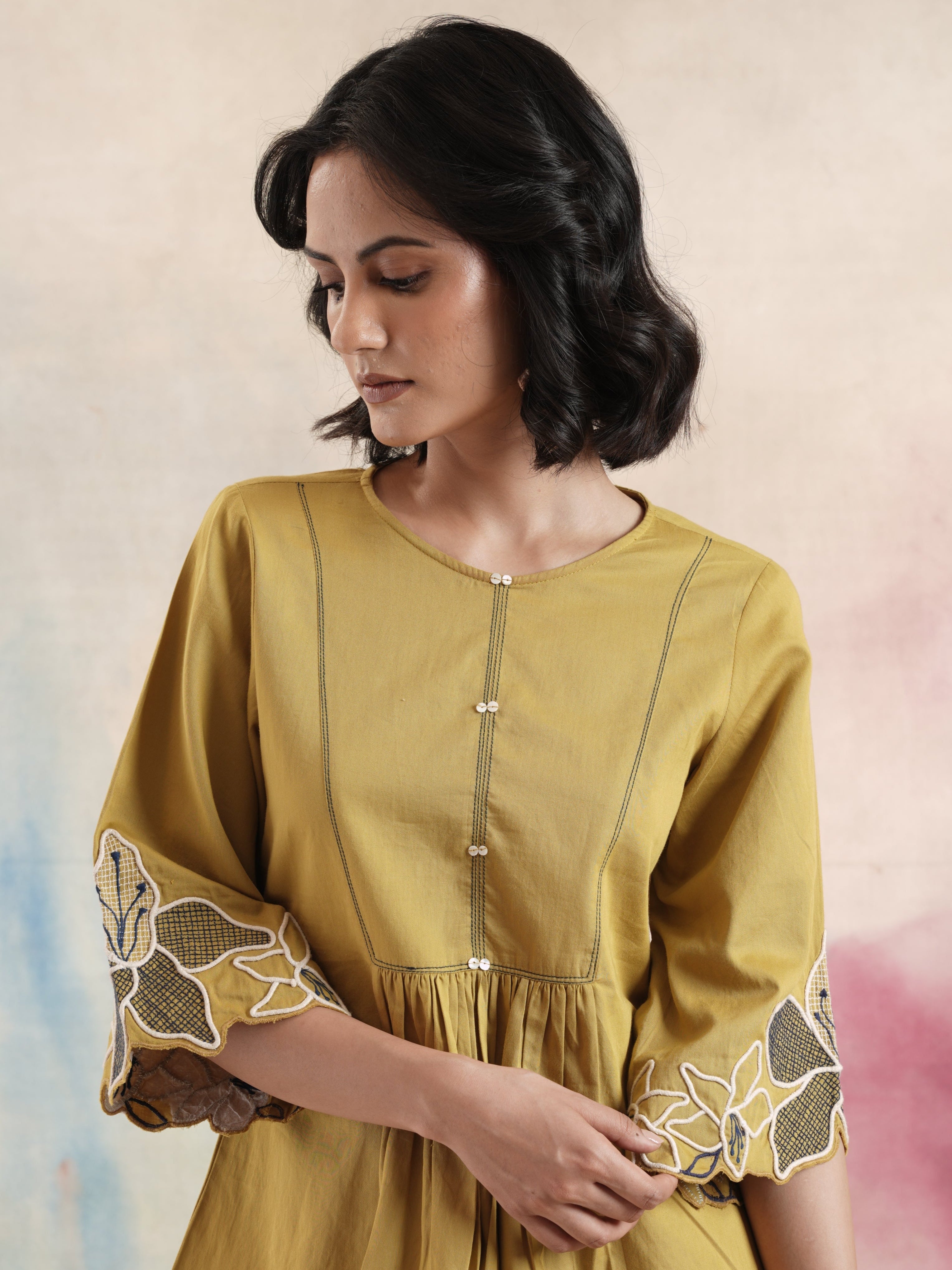 A woman wearing a mustard yellow embroidered tunic with round neckline and 3/4 sleeves, with floral embroidery details.